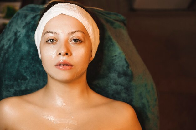 Close up portrait of a beautiful caucasian woman leaning on a spa bed looking at camera while having a anti age mask on her face with hyaluronic acid.