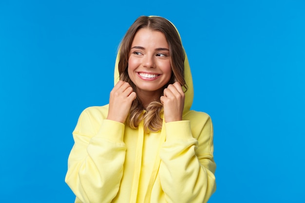 Ritratto del primo piano bella ragazza bionda affascinante messa sul cappuccio della felpa e sorridente, distogliere lo sguardo, sentire freddo come camminare il giorno di primavera, in piedi spensierato