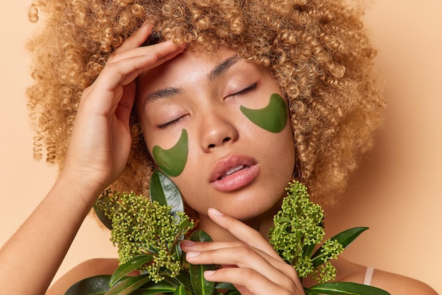 Photo close up portrait of beautiful charming woman keeps eyes closed applies green collagen patches holds branch of fresh plant undergoes beauty treatments has charming look isolated over beige wall