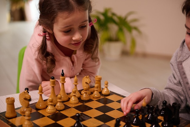 Close-up portrait of a beautiful cute little European girl playing chess game with her elder brother who picks up a chess piece and make his move. Intellectual smart development board games for kids