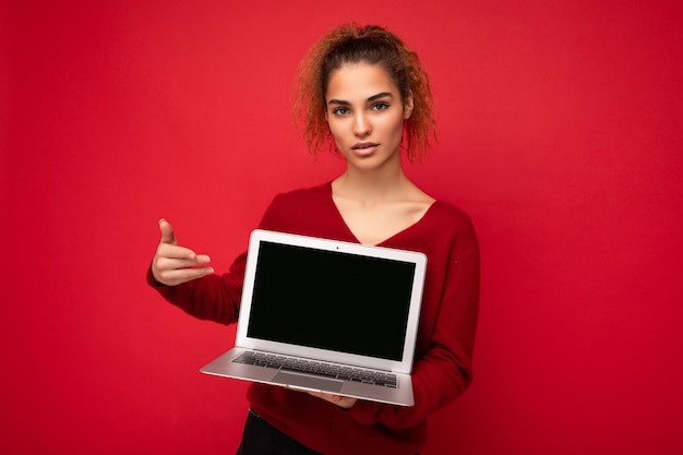 Close-up portrait of beautiful dark blond curly girl holding laptop computer looking at camera pointing finger at netbook monitor wearing red sweater isolated over red wall background.