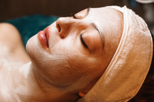 Close up ritratto di un bel viso femminile appoggiato su un letto con gli occhi chiusi e con una maschera bianca sul viso in un salone spa.