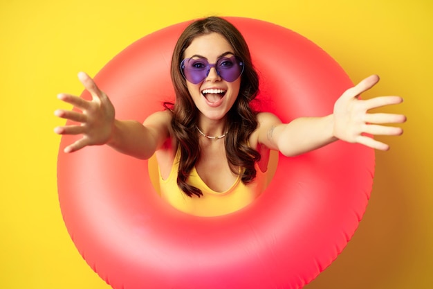 Ritratto ravvicinato di bella donna felice all'interno dell'anello da nuoto rosa, attirando, invitando le persone in vacanza estiva, festa in spiaggia, in piedi su sfondo giallo.