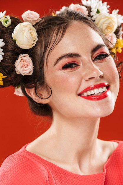 Close up portrait of a beautiful smiling young brunette woman with a floral hairstyle and bright makeup posing isolated over red wall, looking at camera