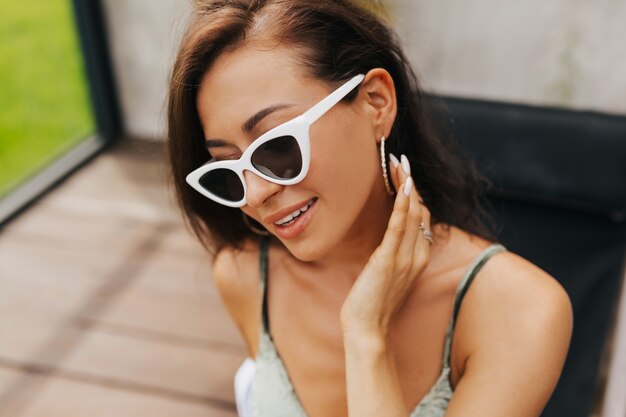 Close-up portrait of beautiful tanned girl in trendy sunglasses lyuing by swimming pool outside. Elegant cheerful young woman  enjoying sun while resting outdoor on summer resort