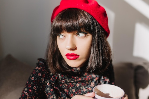 Close-up portrait of beautiful thoughtful girl with red lips and dark hair, looking away while drinking coffee. Attractive french young woman in stylish beret and retro blouse posing during tea time