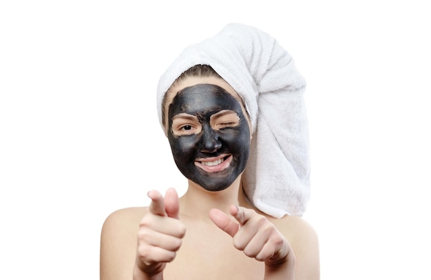 Foto ritratto del primo piano bella donna con maschera facciale nera su sfondo bianco, ragazza con un asciugamano bianco sulla testa, sorriso soddisfatto e felice, indicando le mani verso la telecamera