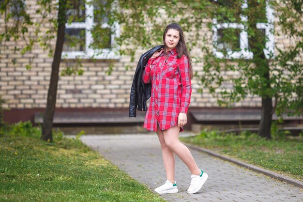 Close up portrait of big plus size beautiful stylish kid girl with leather jacket and red plaid shirt near brick building in urban street as background