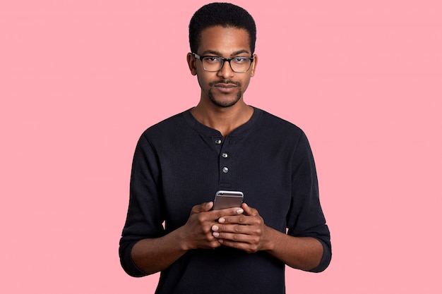 Photo close up portrait of black man with short curly hair, little beard, holds smart phone in hands, writes massage to friends, dressed in casual outfit, isolated on white wall.