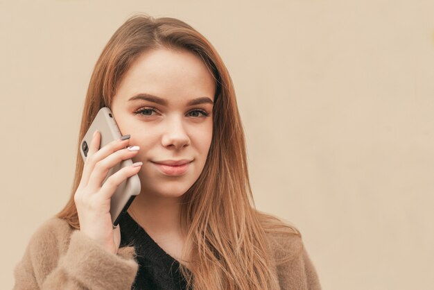 Close-up portrait of a blonde girl calls the phone and looks into the camera on the background of a beige wall, looking into the camera