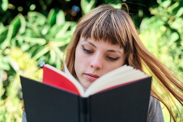 A Close-up portrait of blue-eyed Caucasian blonde young woman reading a book in the park