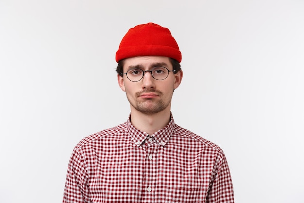 Close-up portrait of bored and reluctant young hipster guy feeling like loser, having bad unlucky day, standing in glasses and red beanie, feel lonely or distressed, stand  