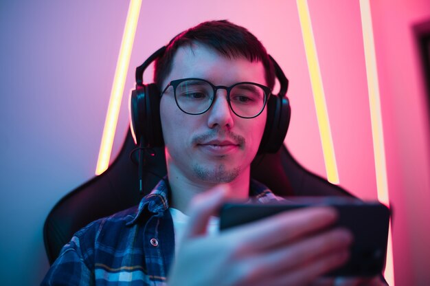 Photo close up portrait of a boy relaxing playing games on mobile phone and sitting in a gaming chair. room with colorful neon light.