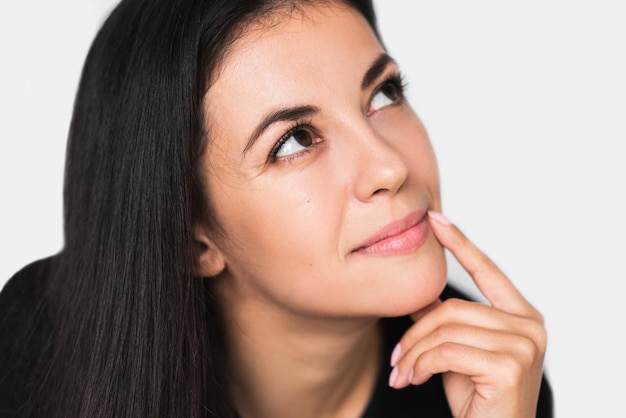 Close up portrait of brunette cute dreamy woman with hand on lips Young pretty woman looking up and wearing black sweater on light grey background Pensive and dreaming mood