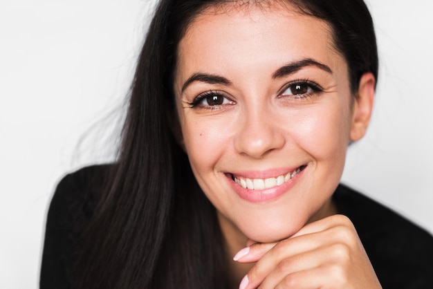 Close up portrait of brunette cute woman with beautiful and healthy toothy smile with hand face Young happy pretty woman wearing black sweater on light grey background Positive and happiness mood