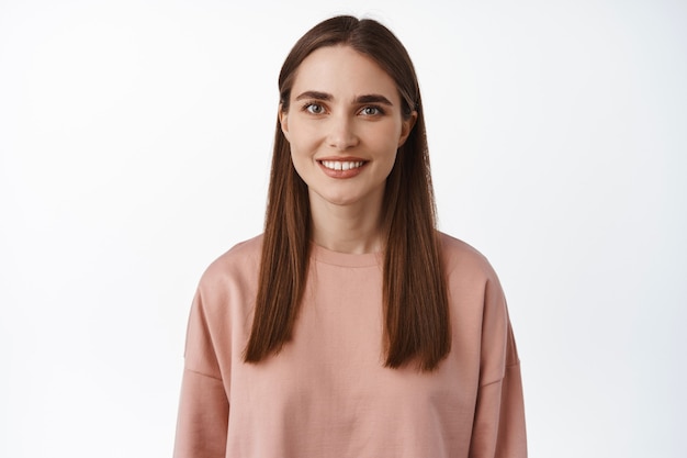 Close up portrait of brunette girl smiling happy on white teeth, looking carefree , standing in pink sweatshirt, casual relaxed pose on white