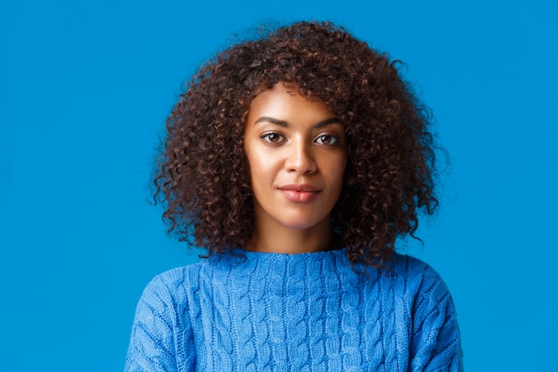 Ritratto di close-up calmo e spensierata bella donna afro-americana in maglione, con acconciatura riccia afro, sorridente carino e fotocamera con espressione amichevole amichevole, blu