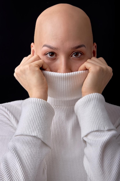 Close-up portrait of caucasian bald female wearing covering half of face with white warm shirt, looking at camera confidently. Isolated over black studio background, copy space. oncology, alopecia