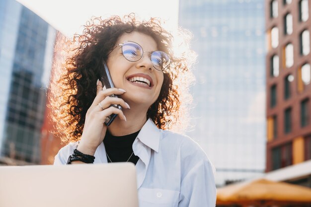 Close up portrait of a caucasian entrepreneur with curly hair and eyeglasses talking on phone while working with a computer outdoor