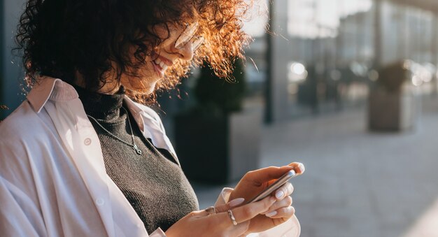 Foto close up ritratto di una donna caucasica con i capelli ricci in chat con qualcuno mentre posa fuori con gli occhiali