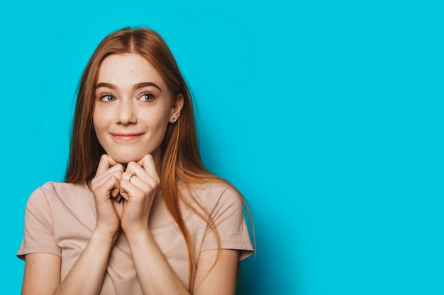 Photo close up portrait of a charming young female looking away thinking while smiling touching face with hands against a blue studio background.