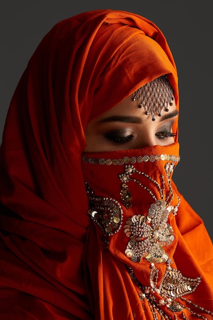 Close-up portrait of a charming young female with beautiful smoky eyes and jewelry on the forehead, wearing the terracotta hijab decorated with sequins. She is posing sideways and looking down on a da