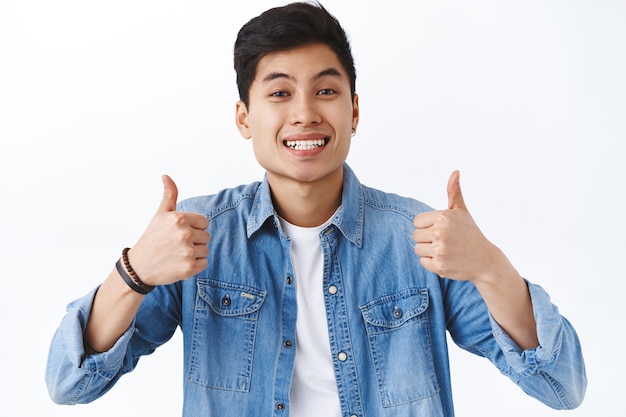 Close-up portrait of cheerful asian young man recommend product or company service, show thumbs-up and smiling, nod in agreement, approve and like idea, white wall