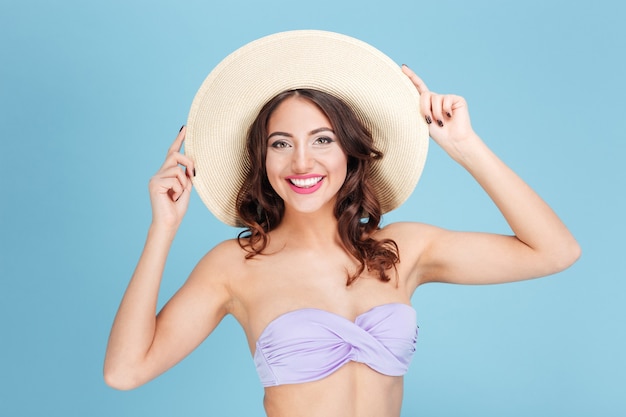 Close-up portrait of a cheerful pretty girl in a beach hat and swimsuit isolated on the blue background