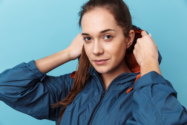 Close up portrait of a cheerful young girl wearing hood standing isolated over blue wall