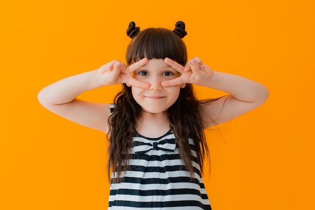 Close up portrait child girl adorable cheerful emotion facial expressing beautiful smile pretty young wearing striped dress isolated yellow wall shows victory fingers, modern kids