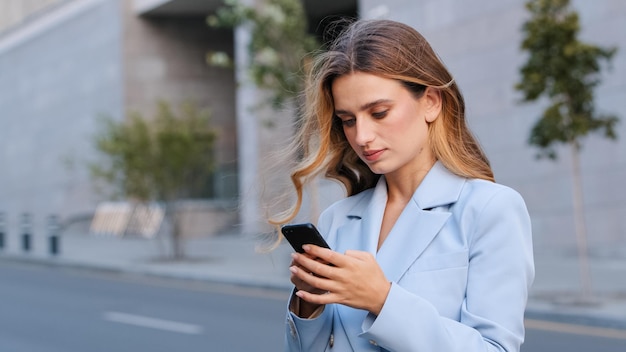 도시 금발의 진지한 여성 비즈니스 여성 사용자의 초상화를 닫고 스마트폰 앱을 사용하여 네트워크 온라인 읽기 뉴스에서 채팅을 위해 휴대 전화 무선 장치 가제트를 보고 거리에 서 있습니다.