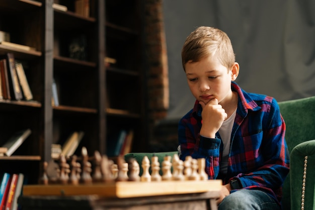 本物のインテリアの部屋の机に座っているあごの近くで手を握ってチェス盤の上で次の動きを考えている賢い物思いにふける小さな男の子のクローズアップの肖像画。チェスをしている就学前の少年。