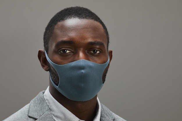 Close up portrait of confident African-American businessman wearing mask and looking at camera while standing against minimal gray background, copy space