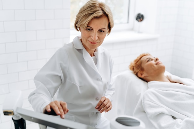 Close up portrait of cosmetologist adjusting settings on monitor for laser hair removal device while young woman lying on couch. Aesthetic body treatment concept