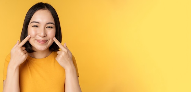 Close up portrait of cute asian girl showing her dimples and smiling coquettish at camera standing over yellow background Copy space