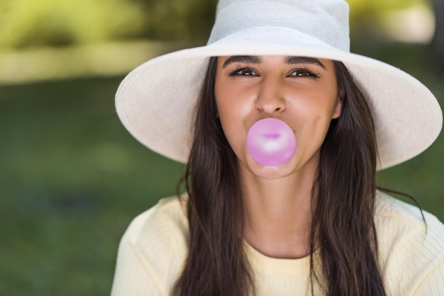 Close up portrait of a cute brunette funny girl in hat chewing bubble gum and looking at camera outdoor on nature background