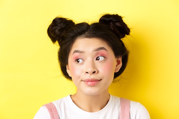 Close-up portrait of cute glamour asian girl, bright makeup and hairstyle, looking aside at logo with silly face, yellow