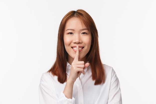 Close-up portrait of cute kind asian girl asking be quiet, keep secret safe, smiling happily and press index finger to lips, looking  make promise, hushing, standing on a white wall