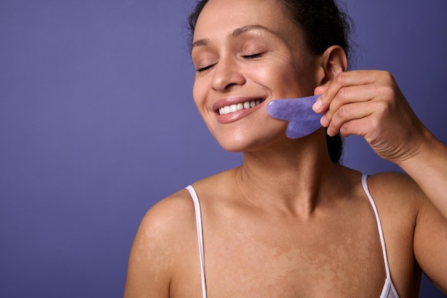 Close-up portrait of delightful self-confident natural beauty\
woman with vitiligo skin problems smiling toothy smile massaging\
her face with ayurvedic gua-sha jade massager against violet\
background