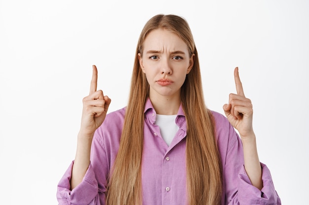 Close up portrait of disappointed female pointing fingers up, grimacing upset and showing bad promo on top, standing upset against white wall