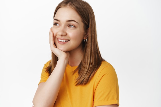 Close up portrait of dreamy happy woman, smiling looking aside with thoughtful daydreaming face, touching clean glowing skin, standing over white wall.