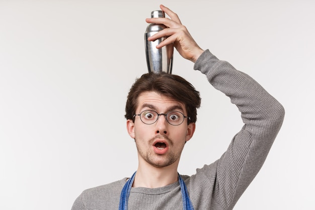 Close-up portrait of enthusiastic funny, handsome male employee, bartender or barista make shocked or surprised expression holding cocktail shaker on head balancing, 