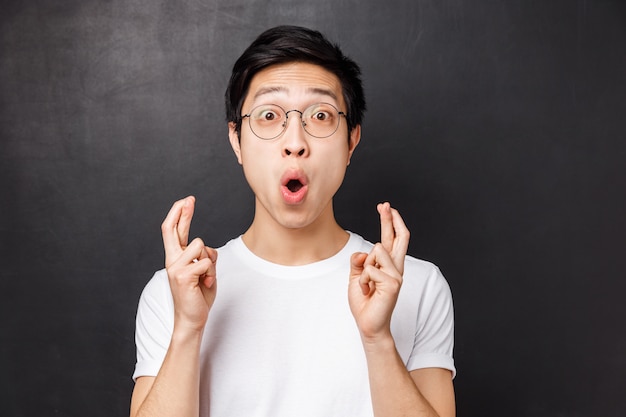 Close-up portrait of excited and hopeful young enthusiastic asian guy in white t-shirt, glasses, cross fingers good luck and staring astounded saying wow, standing black wall amused