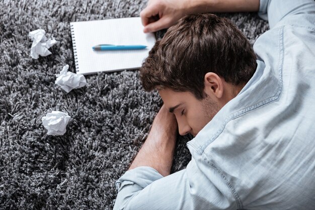 Close up portrait of an exhausted young man sleeping on a carpet with notepad