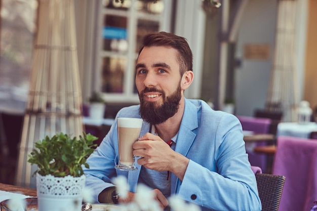 スタイリッシュなヘアカットでファッショナブルなひげを生やした男性のクローズアップの肖像画は、屋外のカフェに座って、カプチーノのグラスを飲みます。