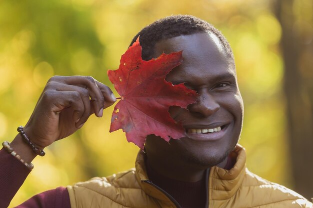 Ritratto ravvicinato di un uomo afroamericano divertente che copre il viso con foglia d'acero autunnale natura autunnale moda autunnale stagionale