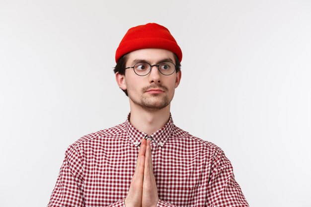 Foto l'uomo caucasico divertente del ritratto del primo piano con i baffi, il berretto rosso e gli occhiali, fa il namaste o prega il gesto distoglie lo sguardo con espressione determinata seria,