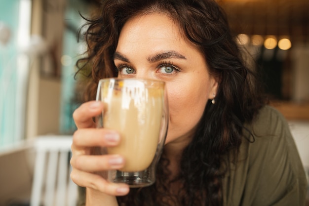 巻き毛の若いきれいな女性の肖像画面白い幸せそうな顔をクローズアップコーヒーショップで飲んでコーヒーを楽しんでいます