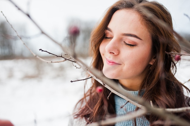 Close up portrait of gentle girl in gray coat near the branches of a snow-covered tree.