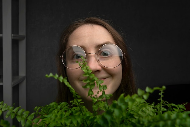 Close-up portrait of girl in round glasses with green houseplant on gray background.